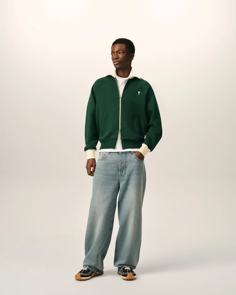 Full-body portrait of a man wearing a green zip-front jacket, white shirt, light blue jeans, and black-and-tan sneakers, standing against a pale background, looking to the side.