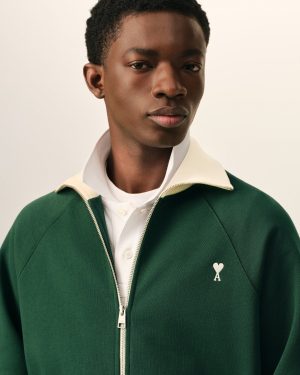 Close-up portrait of a person wearing a green zip-front jacket over a white shirt, against a beige background.