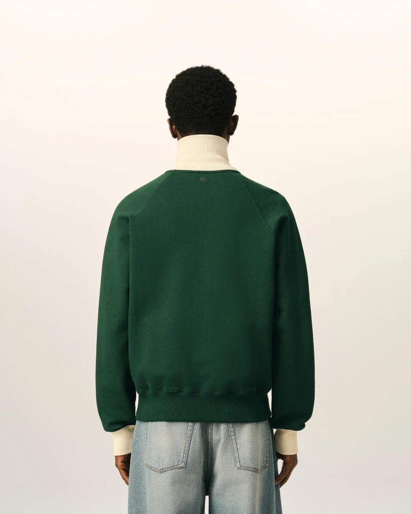 Back view of a person wearing a dark green sweater over a cream turtleneck, with light-wash jeans, standing against a neutral background.