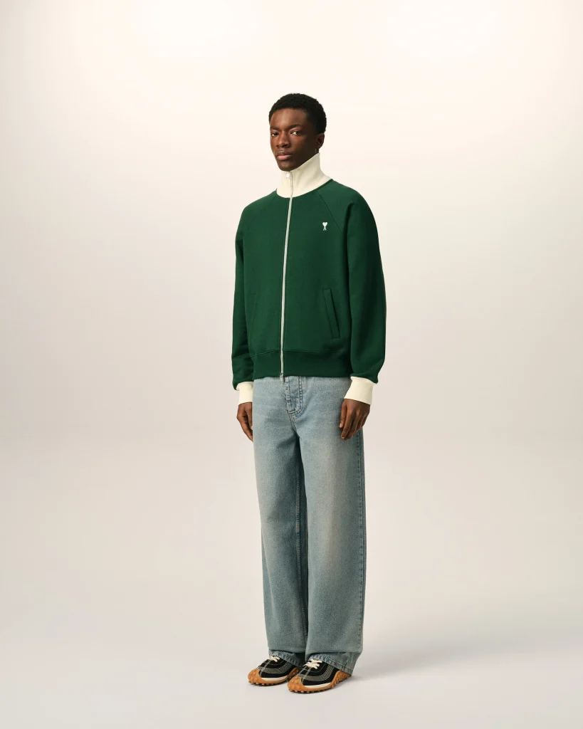 Full-body shot of a man in a green zip-up jacket with a cream collar, light jeans, and patterned sneakers against a neutral backdrop.