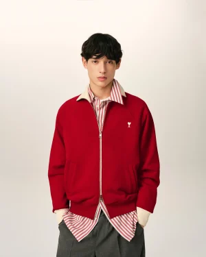 Young man with dark hair wearing a red zip-up jacket over a red-and-white striped shirt, standing against a pale background.