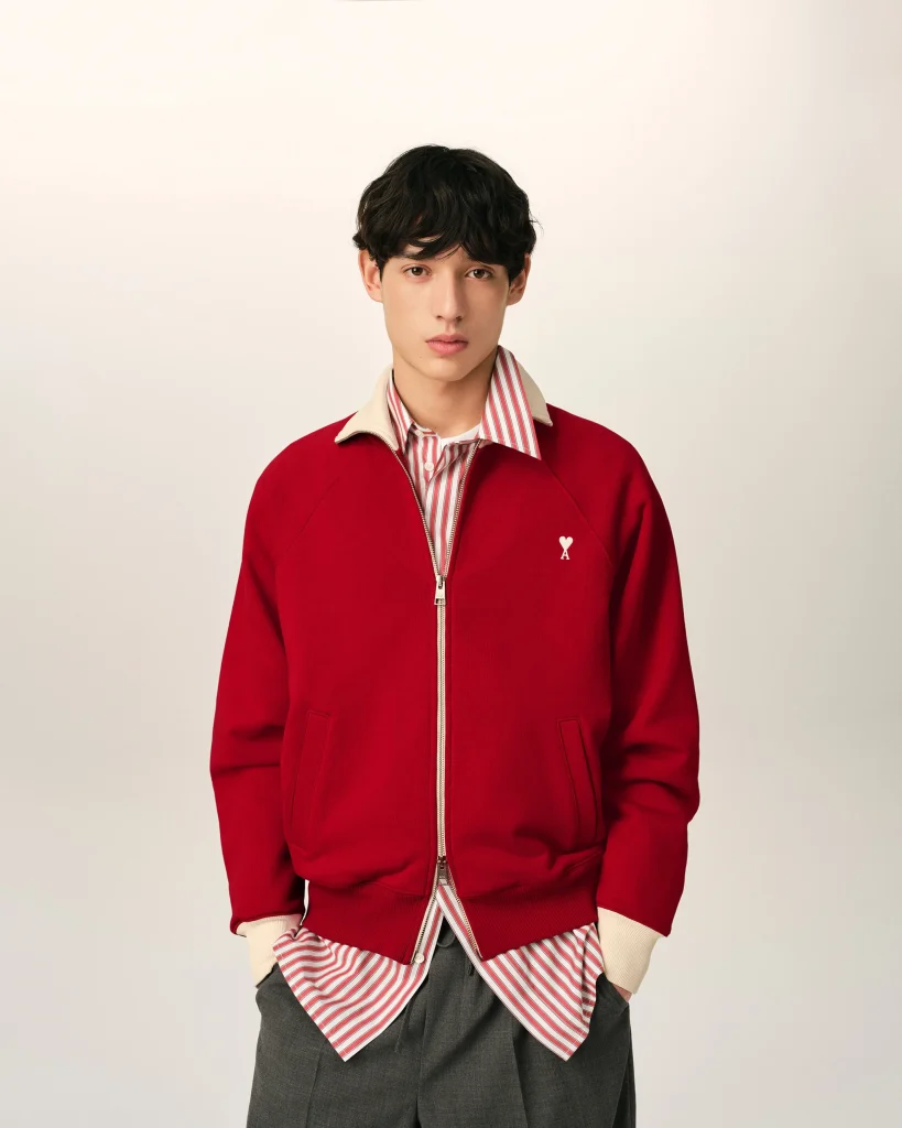 Young man with dark hair wearing a red zip-up jacket over a red-and-white striped shirt, standing against a pale background.