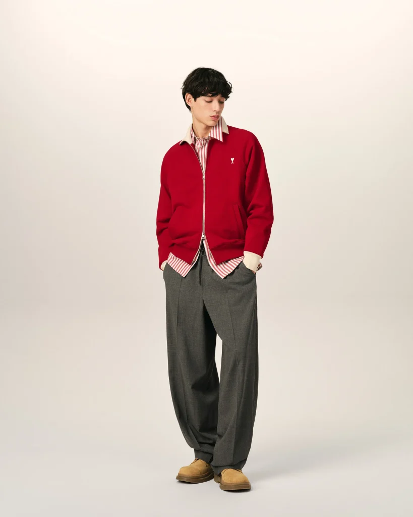 Young man in a red zip-up jacket over a striped shirt, looking downward, standing against a light background.