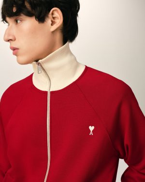 Profile of a person facing left wearing a red zip-front sweater with a beige high collar and a small white heart-and-key emblem on the chest.