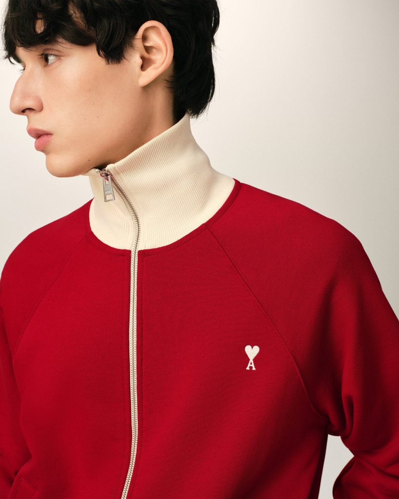 Profile of a person facing left wearing a red zip-front sweater with a beige high collar and a small white heart-and-key emblem on the chest.