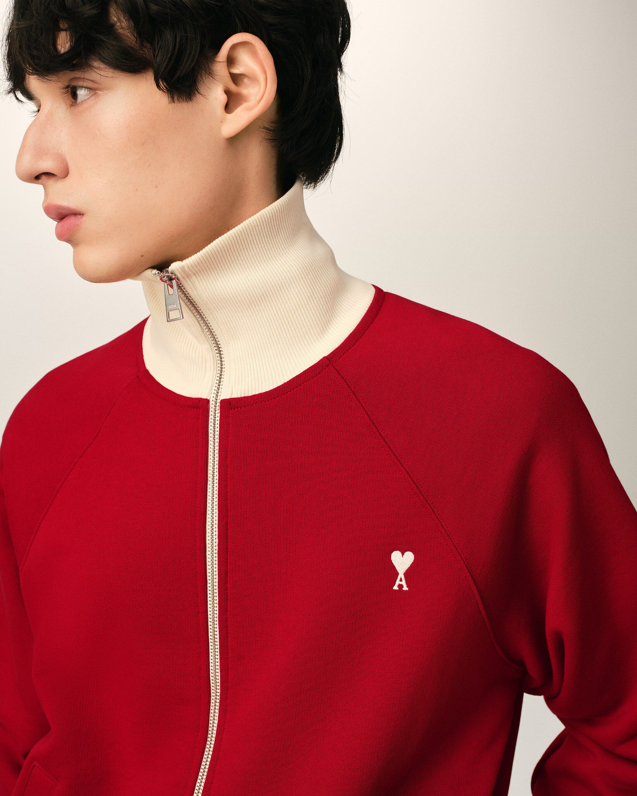 IMG_3451 - New Trend Profile of a person facing left wearing a red zip-front sweater with a beige high collar and a small white heart-and-key emblem on the chest.