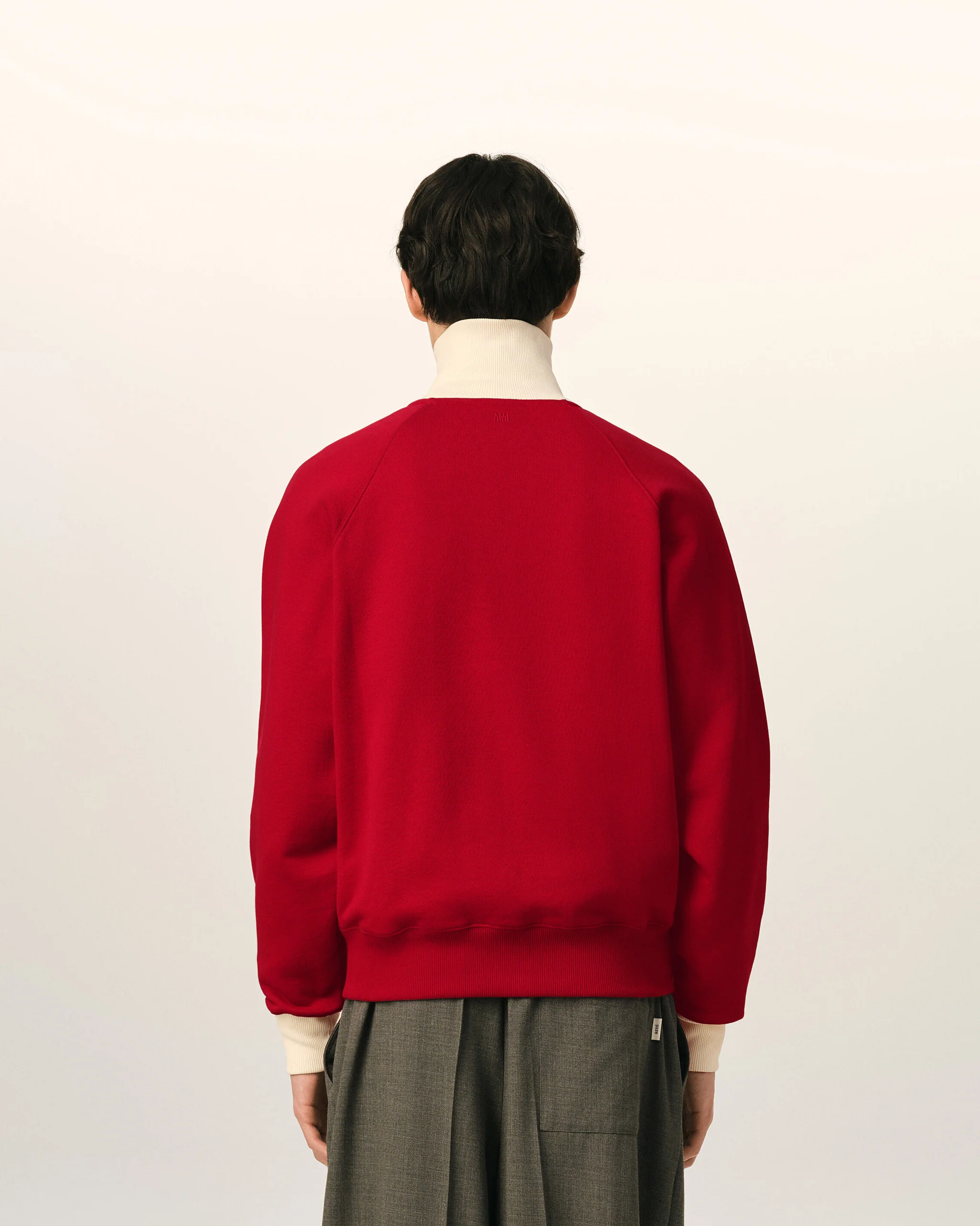 IMG_3452 - New Trend Person standing with back to camera, wearing a red sweater over a white turtleneck and gray pants, against a pale background.