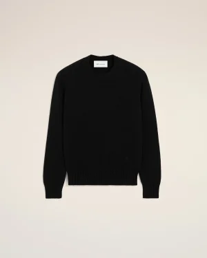 Flat lay of a black knit crewneck sweater on a pale beige background, label visible at the neckline, minimal styling.