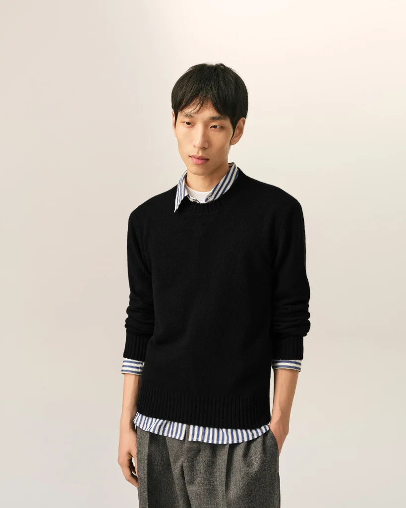 Portrait of a young man wearing a black sweater over a striped collared shirt, looking downward against a light background.