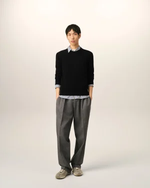 Full-body portrait of a man wearing a black sweater over a striped shirt and gray trousers, standing against a plain beige background.