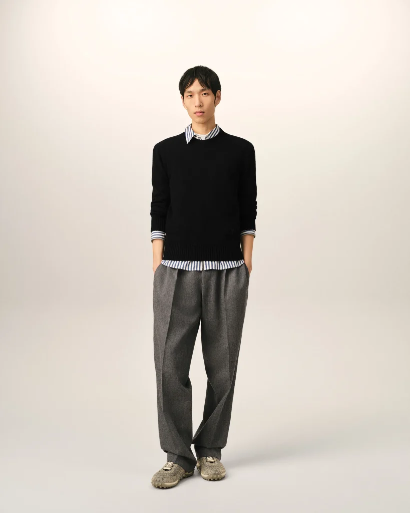 Full-body portrait of a man wearing a black sweater over a striped shirt and gray trousers, standing against a plain beige background.