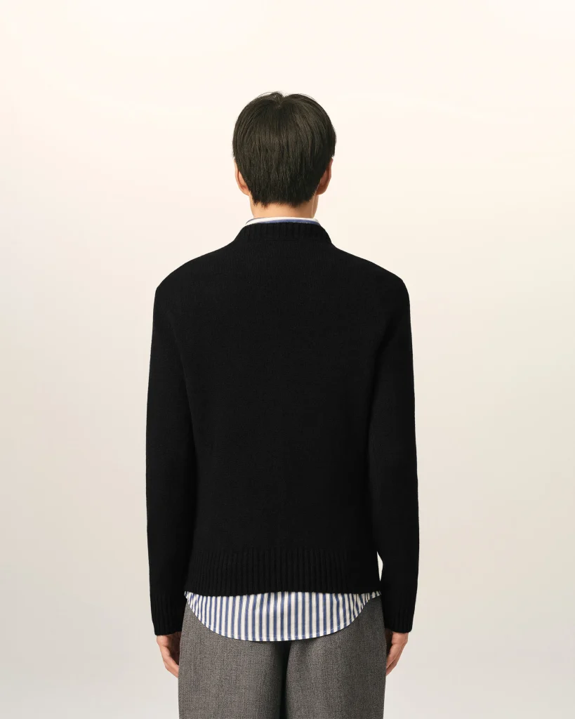 Back view of a person standing, wearing a black sweater over a blue-and-white striped shirt with the shirt hem showing at the bottom and gray trousers visible at the sides.