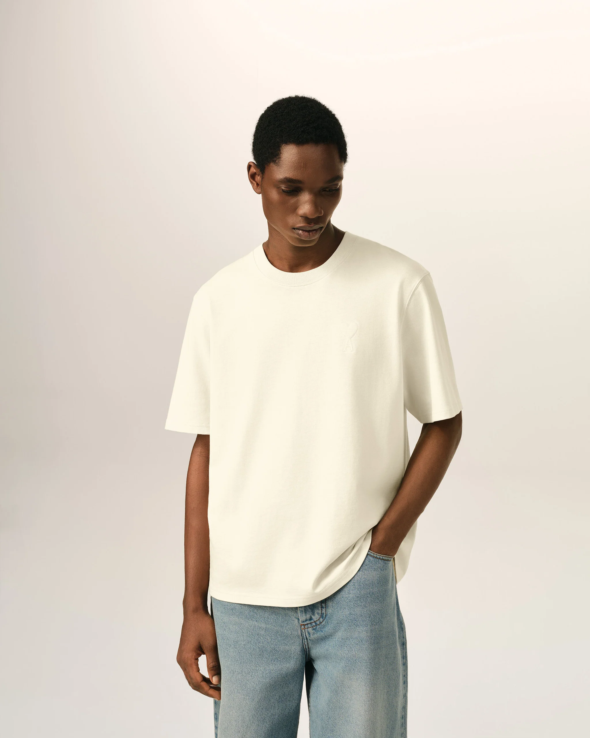 IMG_3460 - New Trend Young Black man wearing a white T-shirt and light-wash jeans, looking down against a pale beige background.