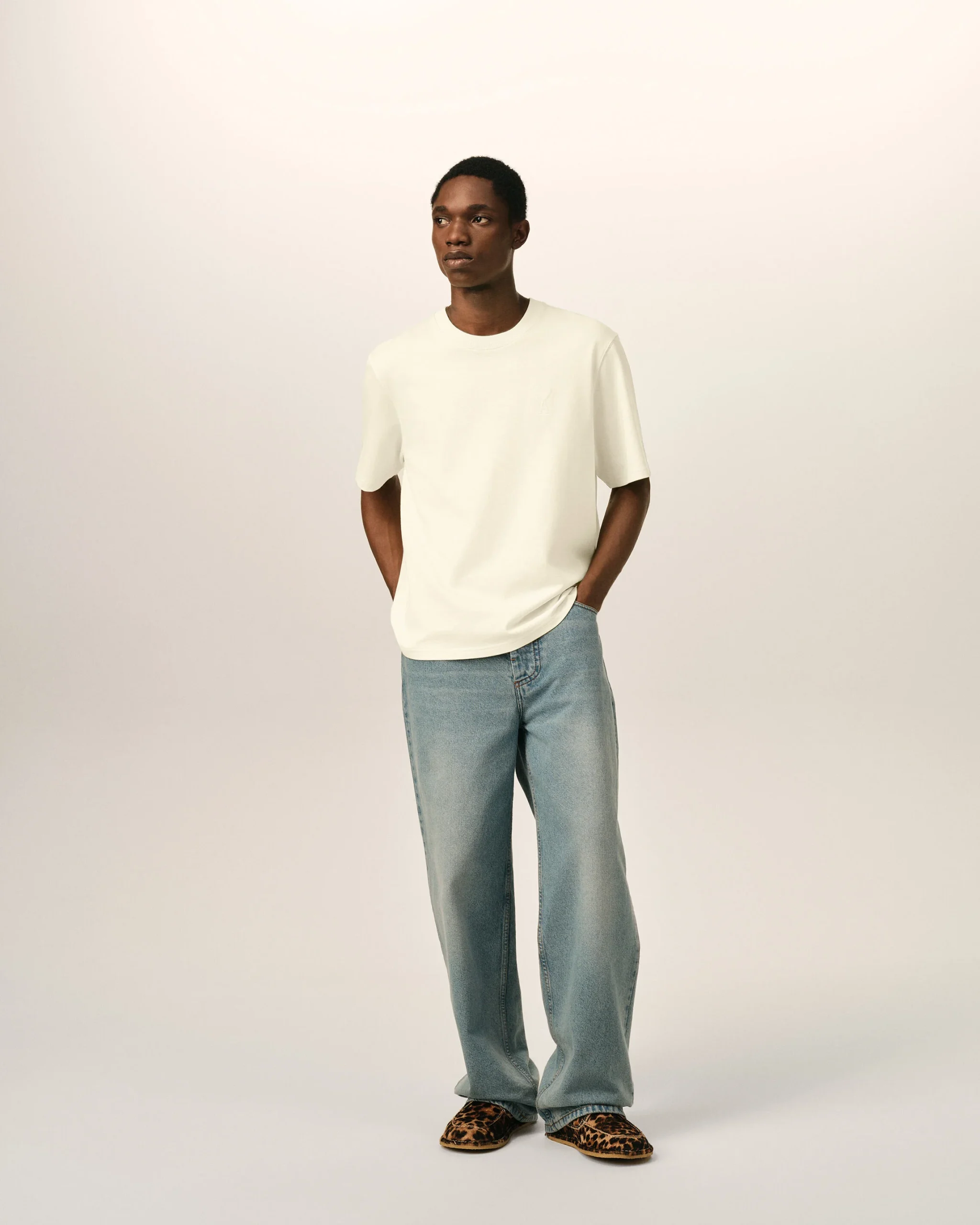IMG_3461 - New Trend Model wearing a white T-shirt and light-wash jeans stands against a neutral beige backdrop, leopard-print shoes visible at the feet.