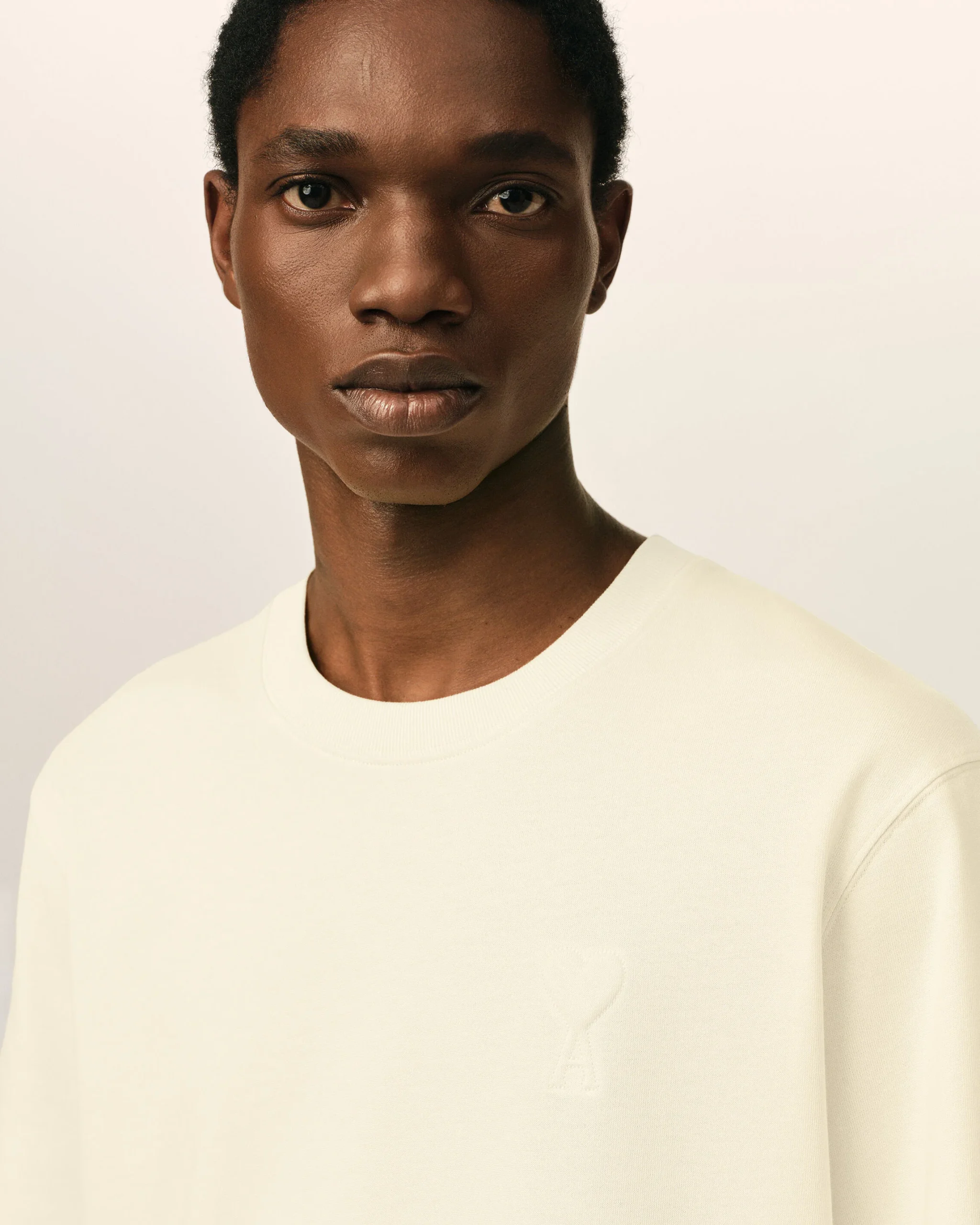 IMG_3462 - New Trend Close-up portrait of a Black person with short hair wearing a white crewneck, against a neutral background, looking at the camera.
