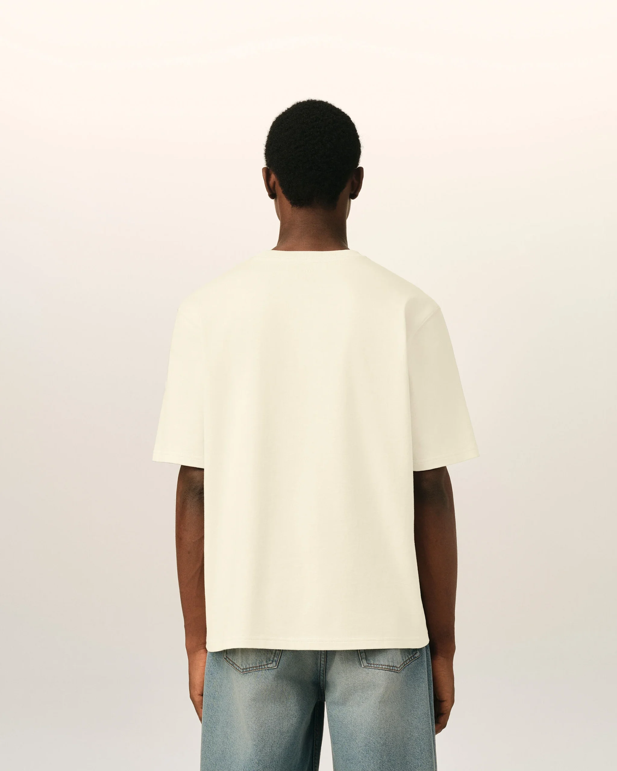 IMG_3463 - New Trend Back view of a person wearing a cream T-shirt and blue jeans against a pale neutral background.