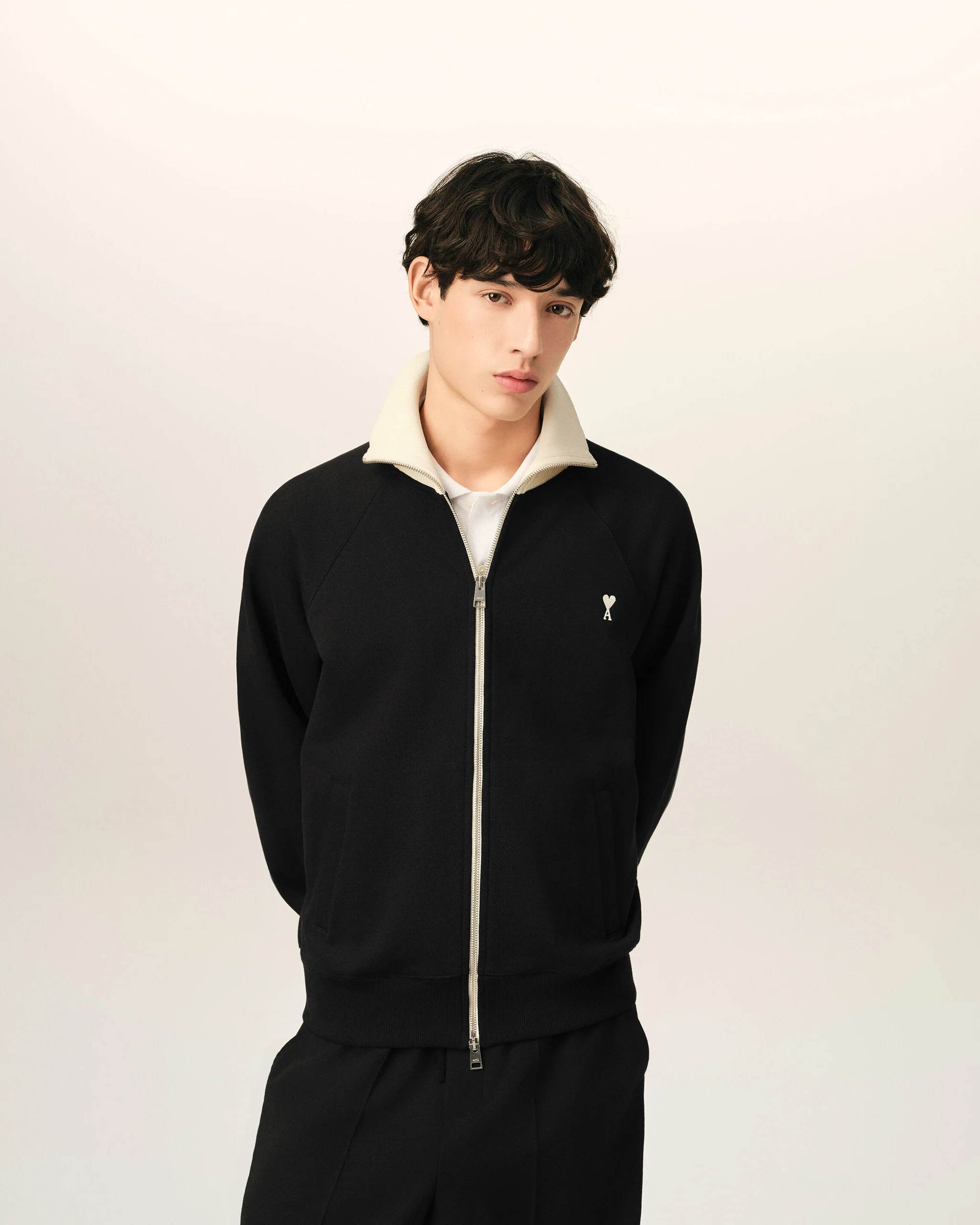 IMG_3471 - New Trend Young man wearing a black zip-up jacket with a cream collar, standing against a light neutral background, facing slightly right with a calm expression.