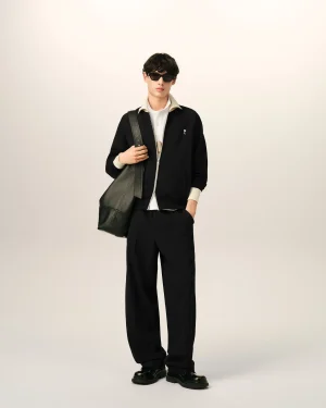 Young man in a black zippered jacket over a white shirt, wearing dark sunglasses and carrying a large green bag over his shoulder.