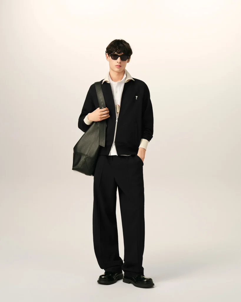Young man in a black zippered jacket over a white shirt, wearing dark sunglasses and carrying a large green bag over his shoulder.