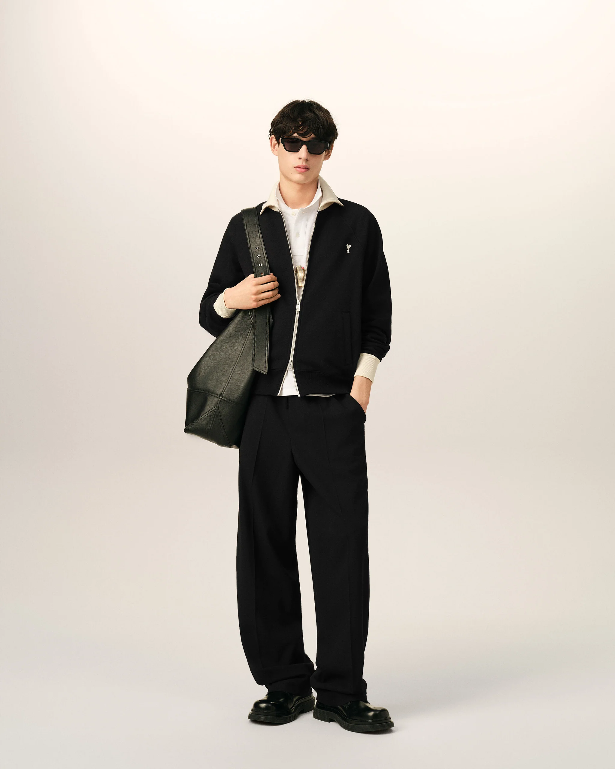 IMG_3472 - New Trend Young man in a black zippered jacket over a white shirt, wearing dark sunglasses and carrying a large green bag over his shoulder.