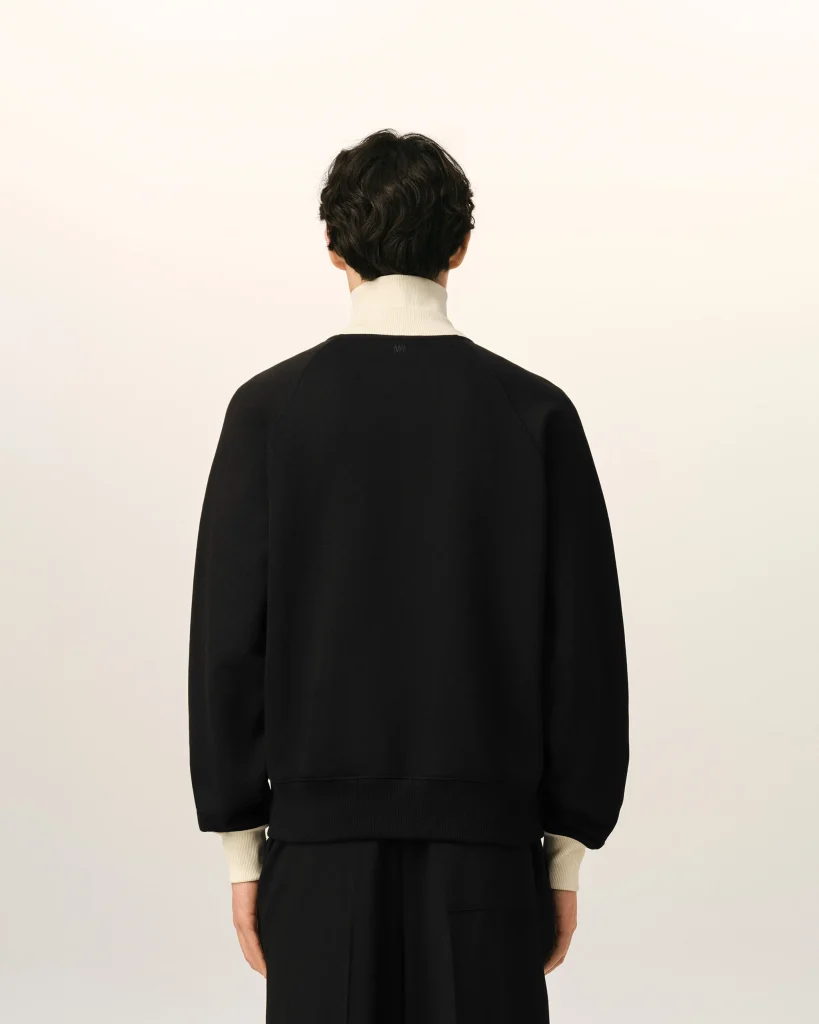 Person shown from behind wearing a black sweatshirt with a light-colored collar and cuffs, standing against a pale wall.