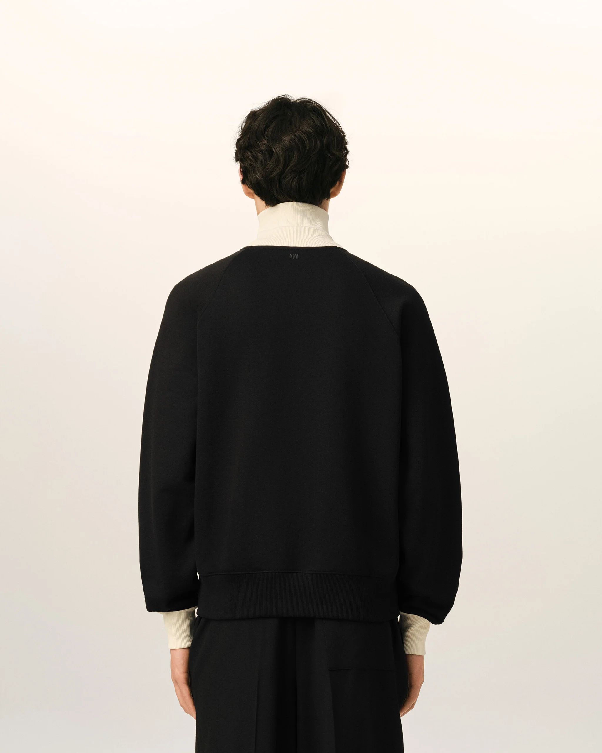 IMG_3474 - New Trend Person shown from behind wearing a black sweatshirt with a light-colored collar and cuffs, standing against a pale wall.
