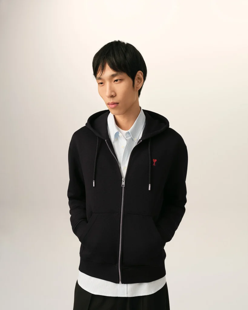 Young man wearing a black zip-up hoodie over a light blue striped shirt, standing against a pale backdrop, looking downward to his left.