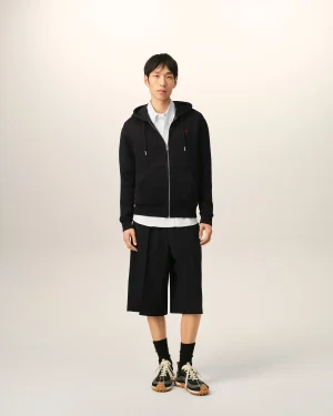 Young man standing and facing the camera wearing a black zip-up hoodie over a light shirt, with wide-leg black shorts and sneakers against a neutral background.