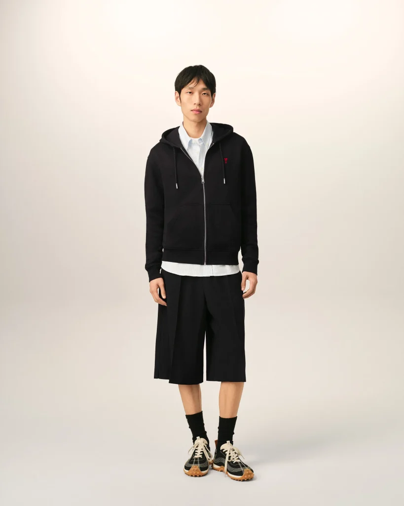 Young man standing and facing the camera wearing a black zip-up hoodie over a light shirt, with wide-leg black shorts and sneakers against a neutral background.