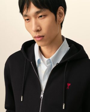 Close-up of a young man wearing a black zip hoodie over a light blue striped shirt, with a red ribbon embroidery on the chest.