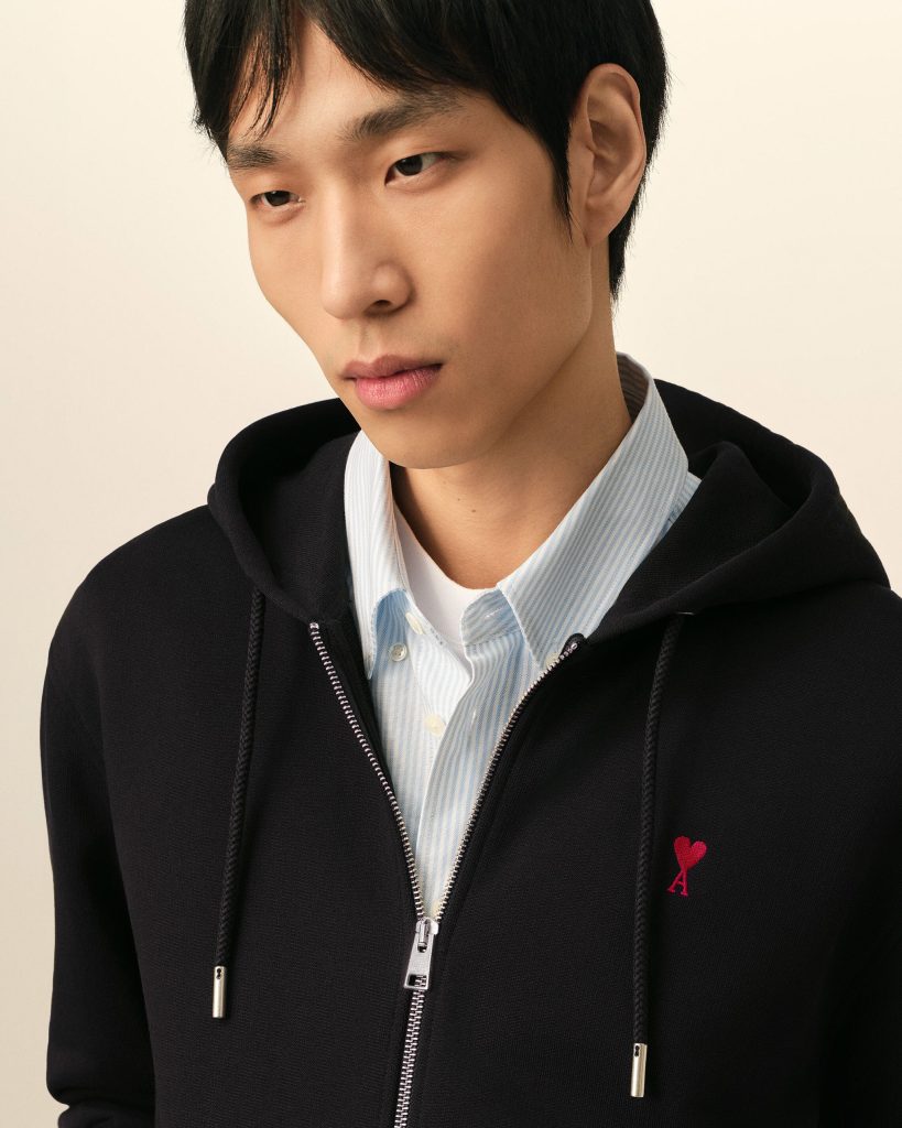 Close-up of a young man wearing a black zip hoodie over a light blue striped shirt, with a red ribbon embroidery on the chest.