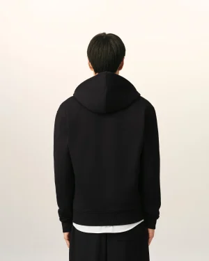 Back view of a person wearing a black hoodie against a light beige background; white shirt hem peeks below the hoodie edge.
