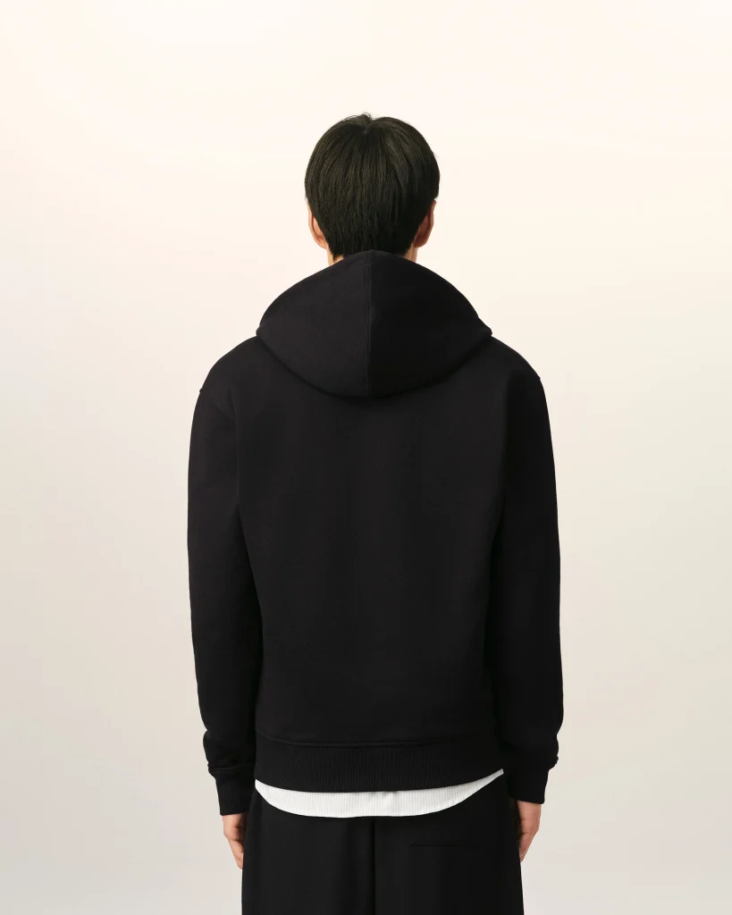 Back view of a person wearing a black hoodie against a light beige background; white shirt hem peeks below the hoodie edge.