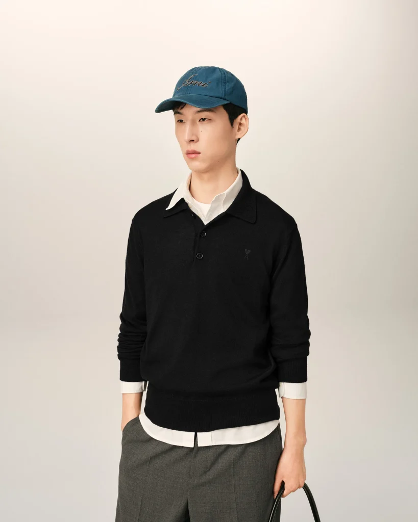 Person wearing a blue cap, black knit sweater over a white shirt, standing against a light neutral background, holding a bag.