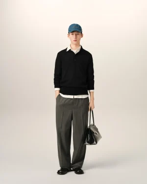 Full-body portrait of a man standing against a pale background, wearing a blue cap, black sweater, white shirt, gray trousers, and holding a black bag.