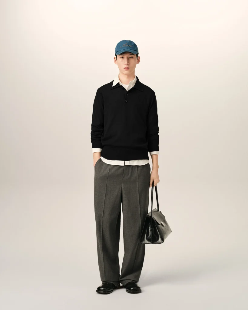 Full-body portrait of a man standing against a pale background, wearing a blue cap, black sweater, white shirt, gray trousers, and holding a black bag.