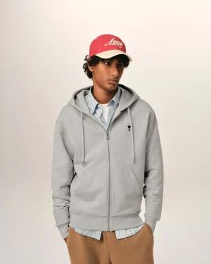 Young person wearing a red cap, grey hoodie, and striped shirt, standing against a light background, looking to the side.