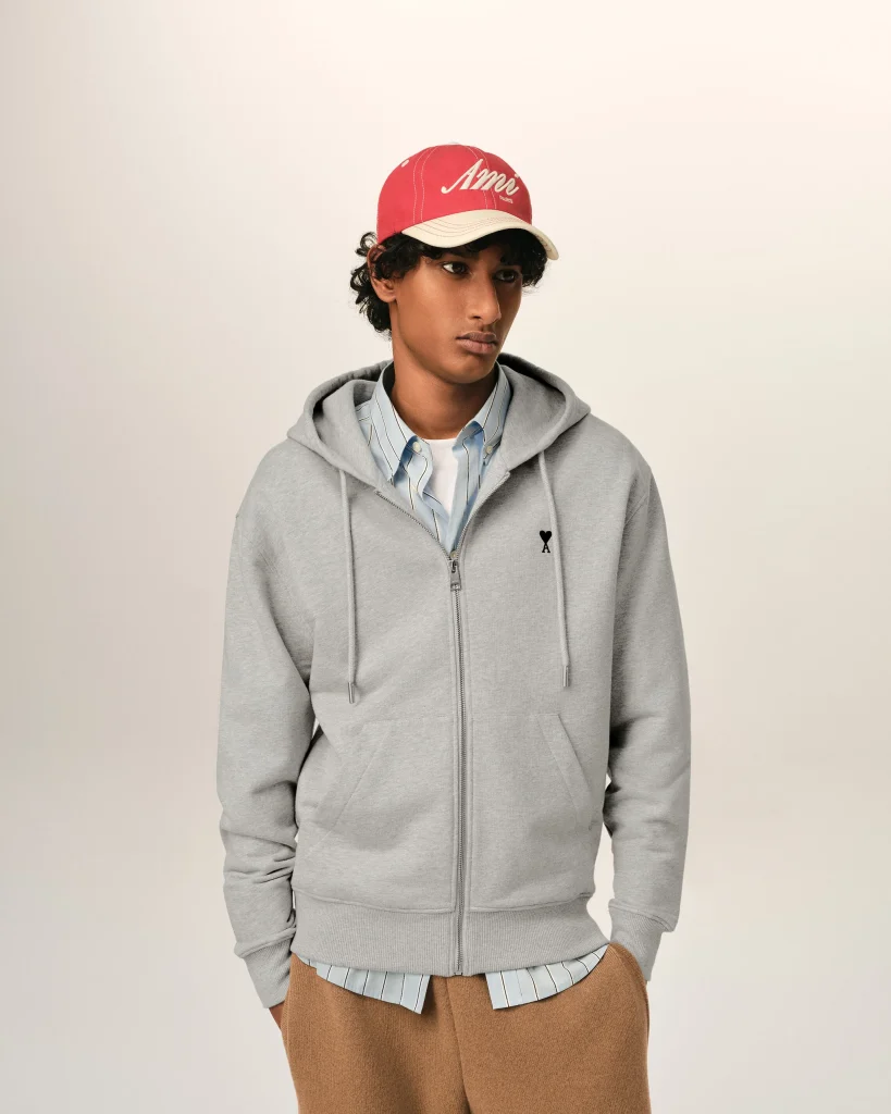 Young person wearing a red cap, grey hoodie, and striped shirt, standing against a light background, looking to the side.