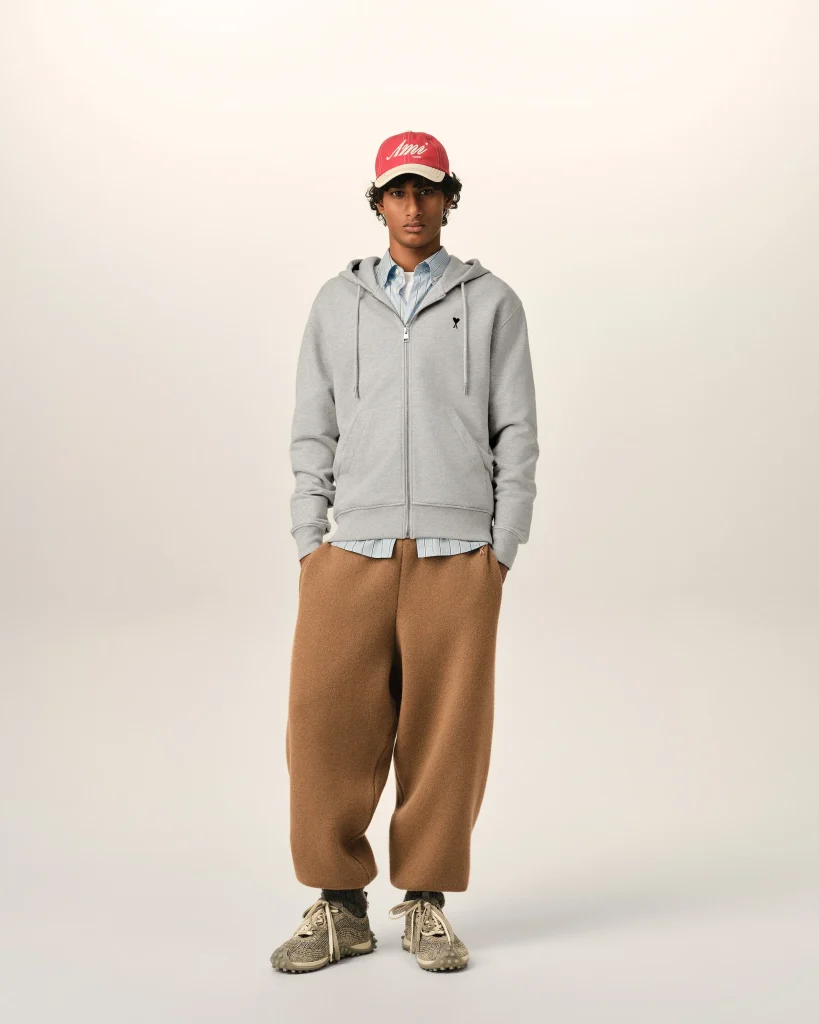 Front view of a person wearing a gray zip hoodie over a blue striped shirt, tan wide pants, gray sneakers, and a red cap, with hands in pockets.