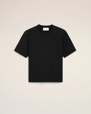 Flat-lay of a black short-sleeve t-shirt on a beige background; small stitched heart logo on the chest and a white neck tag visible.