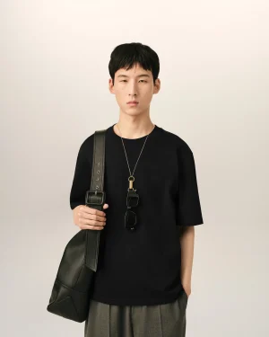 Young person standing against a pale backdrop wearing a black shirt, crossbody bag, and a pendant with sunglasses hanging from the strap