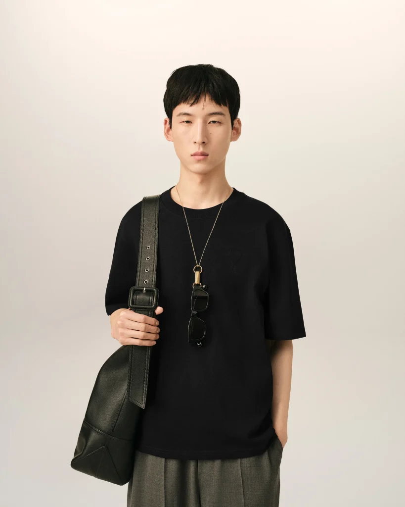 Young person standing against a pale backdrop wearing a black shirt, crossbody bag, and a pendant with sunglasses hanging from the strap