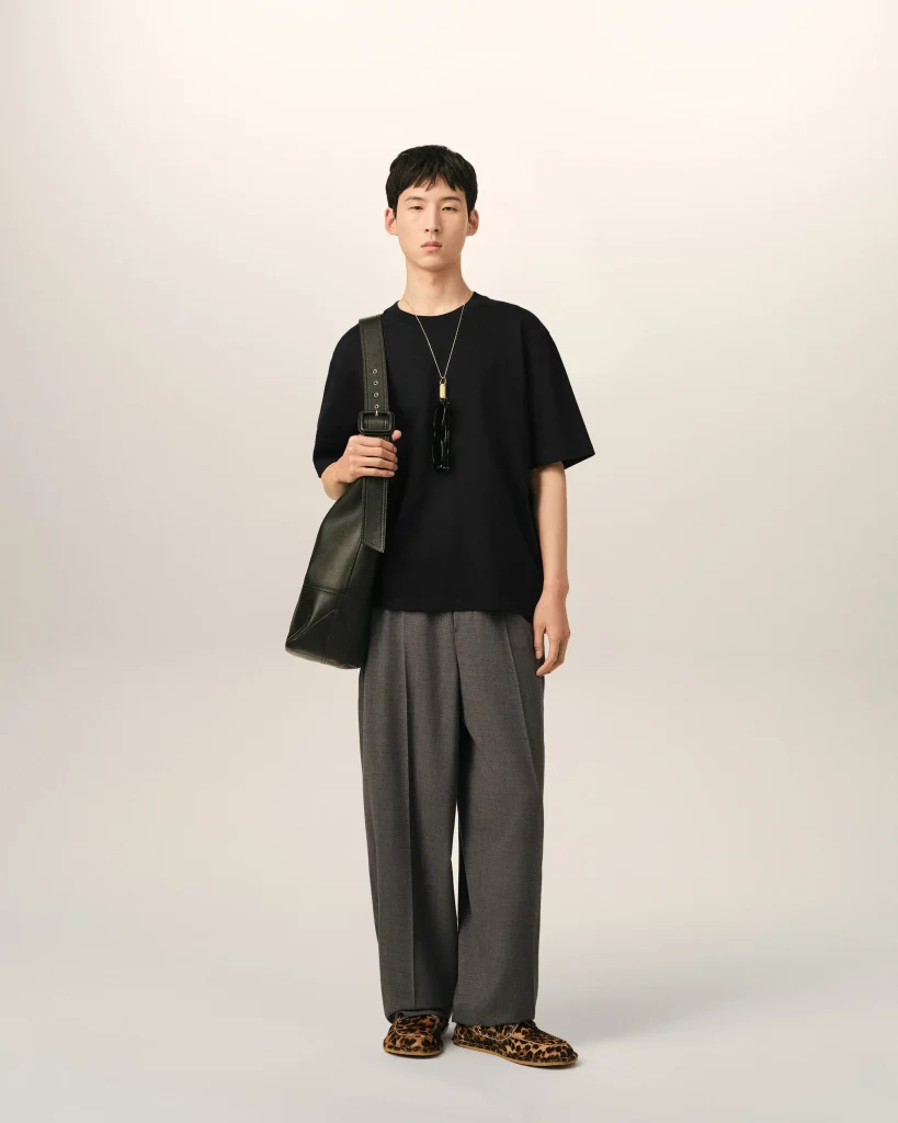 Young person standing against a pale background, wearing a black T-shirt, gray pleated wide-leg pants, leopard-print shoes, and carrying a dark shoulder bag with sunglasses hanging from a necklace.