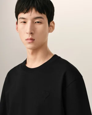Close-up of a person with short black hair wearing a black crew-neck shirt; a small heart embroidery on the chest.