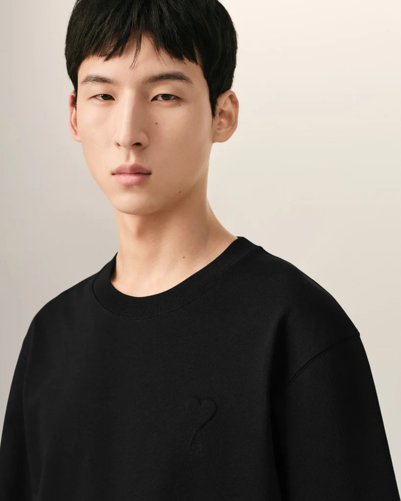 Close-up of a person with short black hair wearing a black crew-neck shirt; a small heart embroidery on the chest.