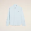 Light blue striped button-down shirt with a small black heart emblem on the left chest against a beige background.