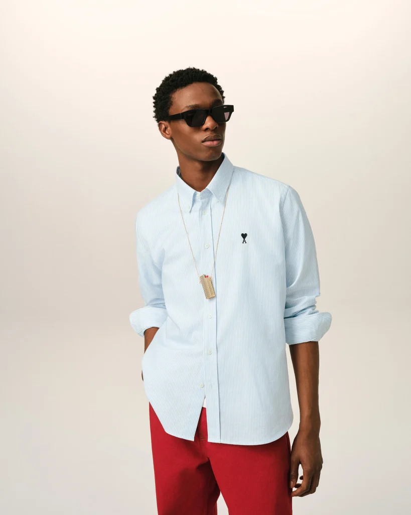 Young man wearing a light blue striped button-down shirt, black sunglasses, and red pants, posing against a pale background.