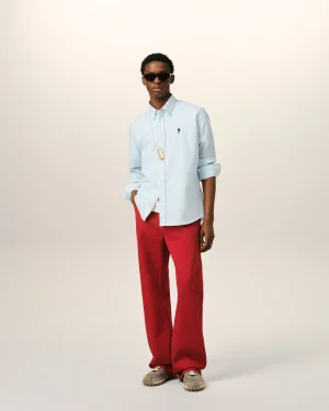 Man wearing a light blue button-down shirt, red pants, and sneakers, posing against a pale background with dark sunglasses and a necklace.