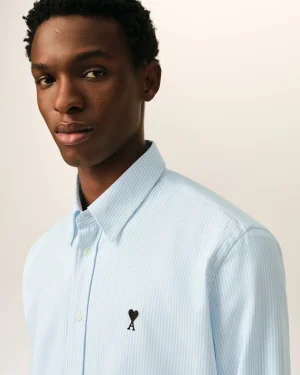 Close-up portrait of a person with short curly hair wearing a light blue striped shirt that has a small black heart and the letter A embroidered on the chest area.