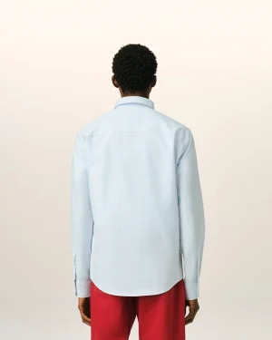 Person seen from behind wearing a light blue striped shirt and red pants against a pale wall backdrop.